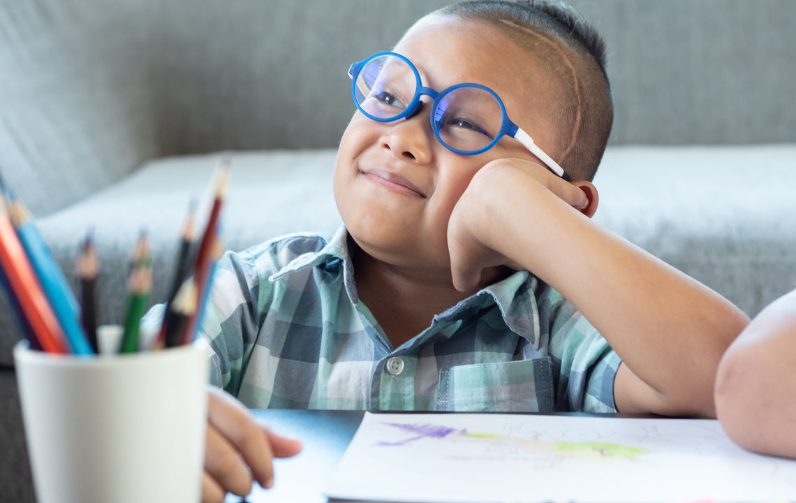 A young boy wearing glasses colors at a table.