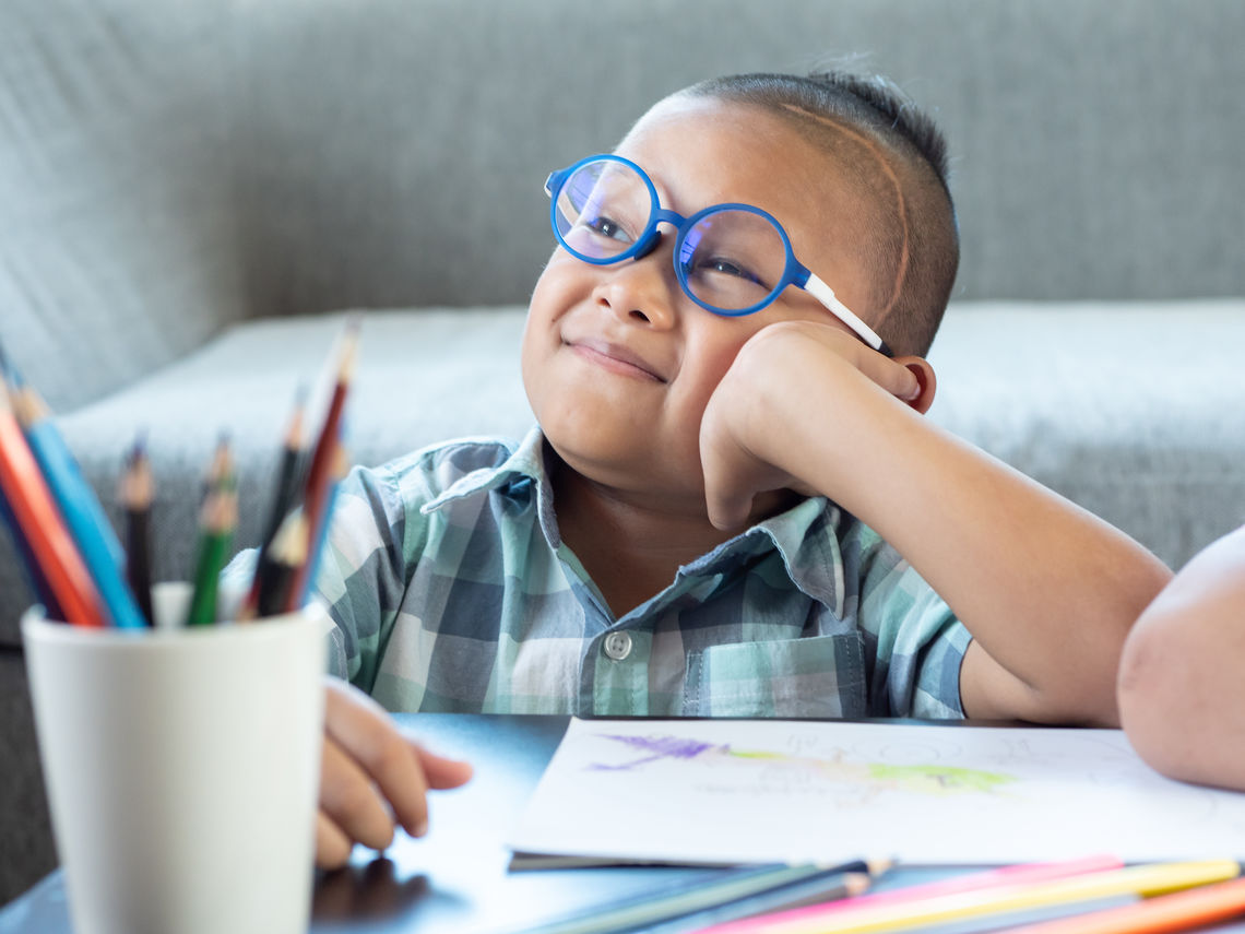 A young boy wearing glasses colors at a table.