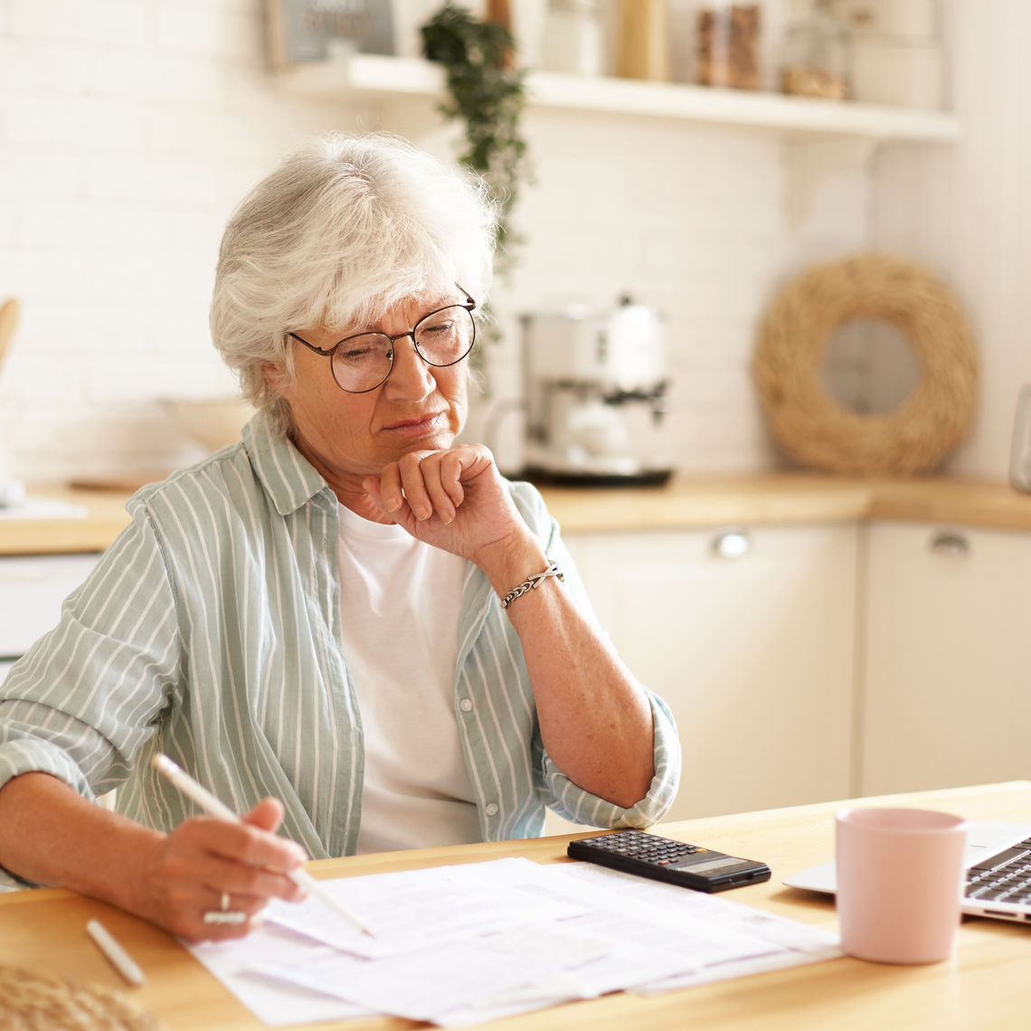 Casually dressed gray haired woman on retirement managing family budget at home, sitting in kitchen with bills, laptop, calculator and mug, writing down in papers using pencil, having focused look