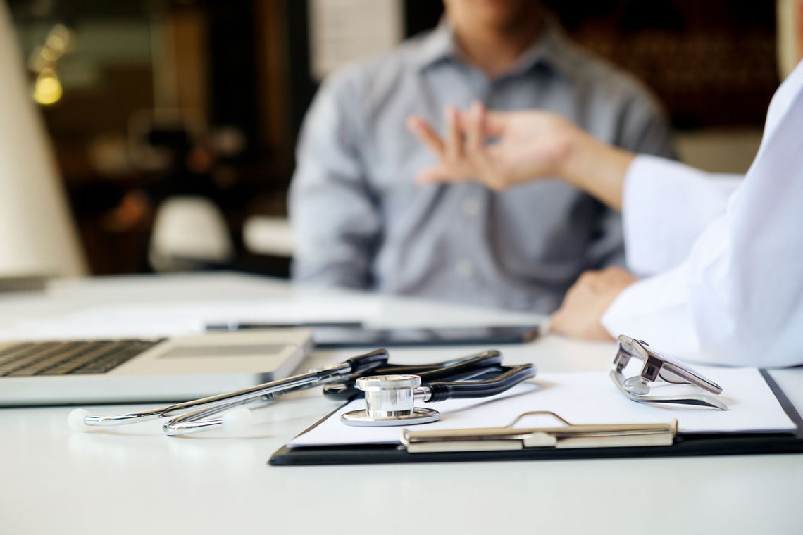 Two doctors have a discussion; they are seated at a desk with a stethoscope, laptop, and clipboard.