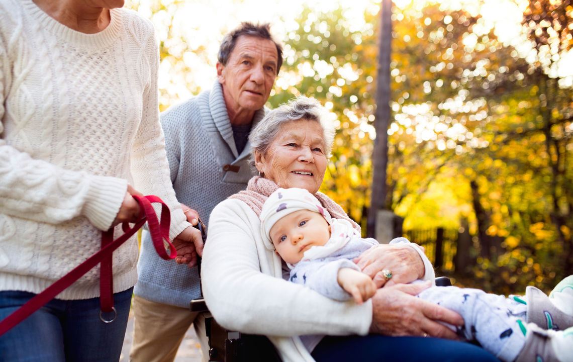 Senior couple with a dog and elderly woman in wheelchair holding a baby. An extended family on a walk in autumn nature.