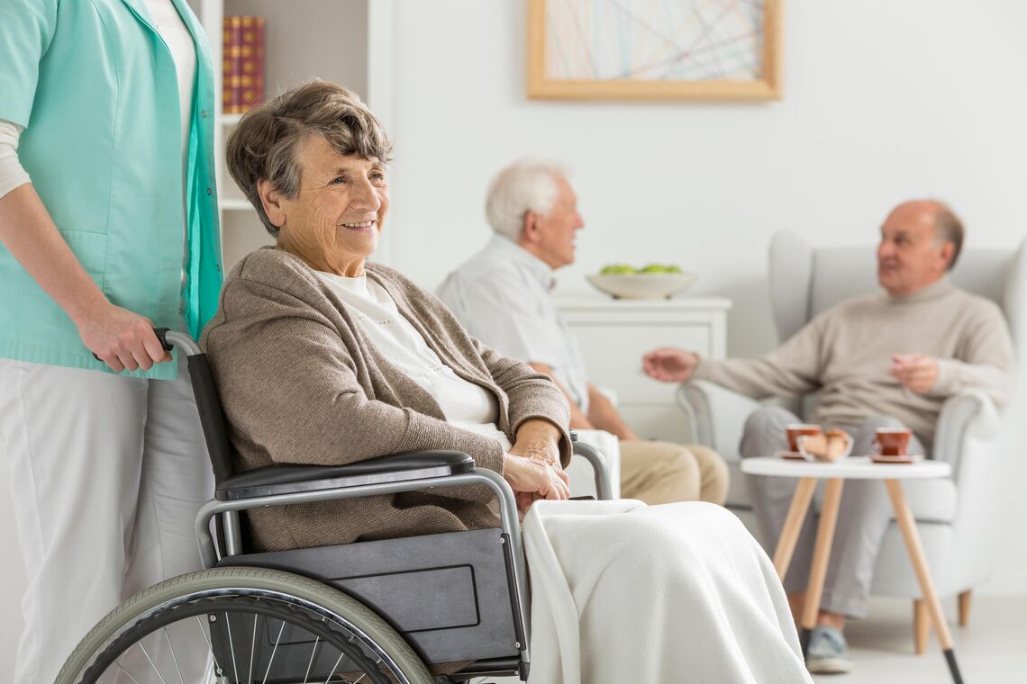 A senior woman is pushed in a wheelchair by an attendant. Two senior men are seated having a conversation in the background.