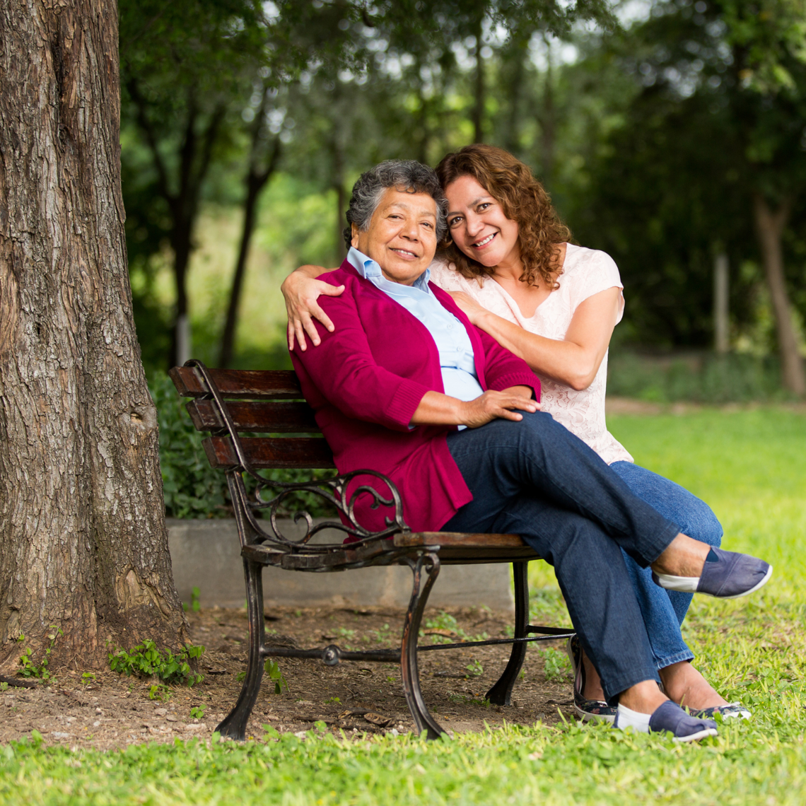 A senior woman and a middle aged woman embrace on a bench under a tree.