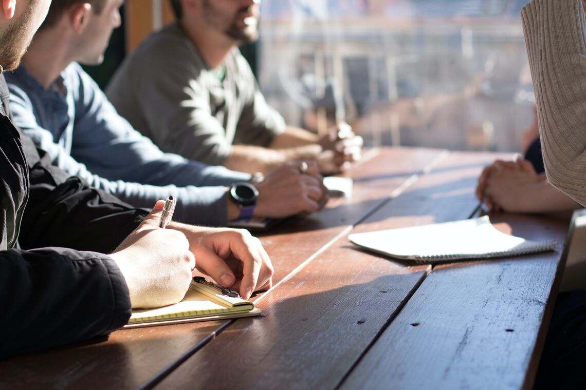 A group of people have a meeting at a table; one person is standing up and talking.