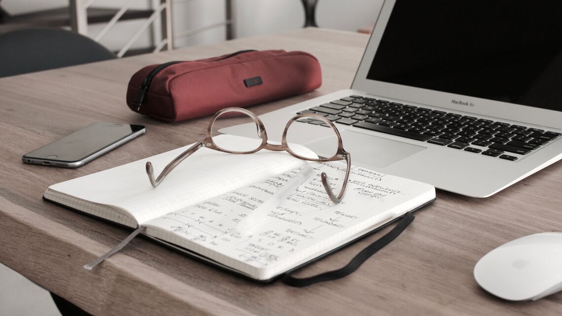 A laptop, notebook, glasses, pencil case, and phone sit on a desk.