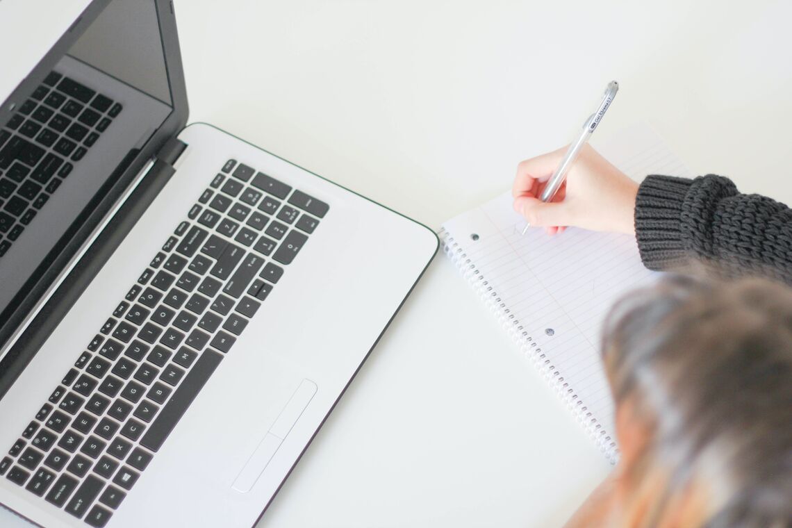 A woman sits at a table in front of a laptop and writes in a notebook.