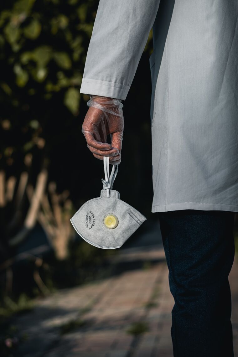 A man in a white coat holds an N-95 mask in his gloved hand.