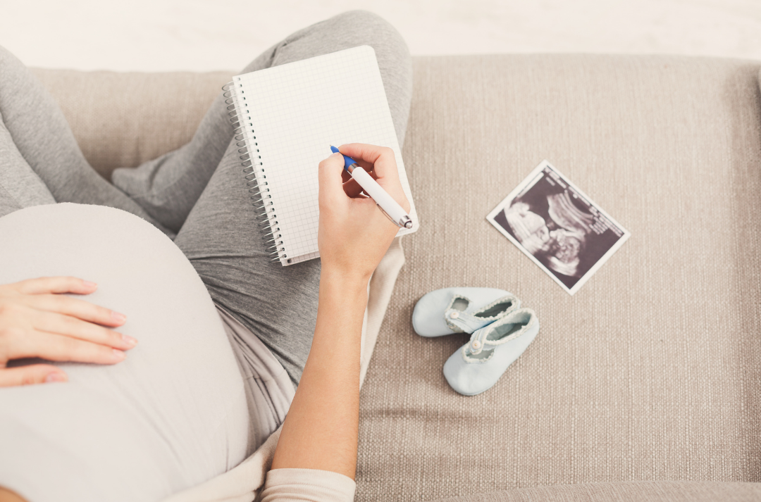 A pregnant person sits on a sofa, with one hand on their stomach and the other writing in a notebook. A pair of baby booties and an ultrasound photo are next to them on the sofa.